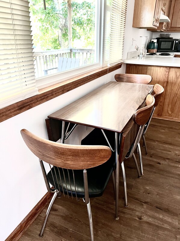 A dining area with a wooden table and chairs, a window with blinds, and a kitchen area with a microwave. - Spring River Waterfront Cabin - Walking Distance to Downtown Hardy & Loberg Park (Hardy)