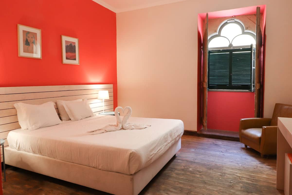 A room with a bed, a chair, a window with shutters, and two framed pictures on the wall.