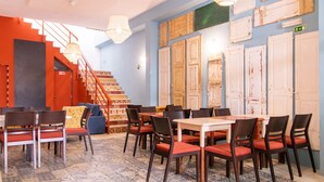 A dining area with tables and chairs, a staircase, and a wall with doors and a green exit sign. - Tilia Hostel (Faro)