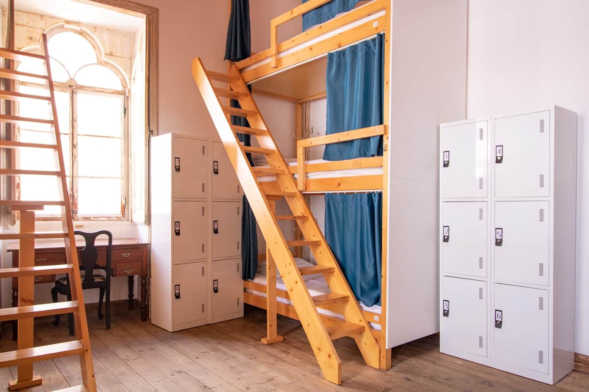 A dormitory room with bunk beds, a wooden ladder, a desk, and storage lockers.