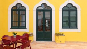 A yellow building with three arched windows and a central door. There is a red table and chairs in front of the building. - Tilia Hostel (Faro)