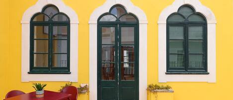 A yellow building with three arched windows and a central door. There is a red table and chairs in front of the building.