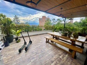 An outdoor patio with a wooden table and benches, surrounded by potted plants and two electric scooters. - Luxury Apartment in Poblado (medellin)