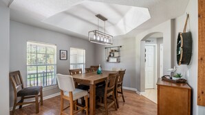 A dining area with a wooden table and chairs, a hanging light fixture, and a view of the outdoors through large windows.