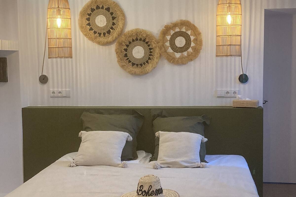A neatly made bed with a white bedspread, two pillows, and a woven headboard. There are three wall-mounted lamps and two decorative woven wall hangings above the bed.