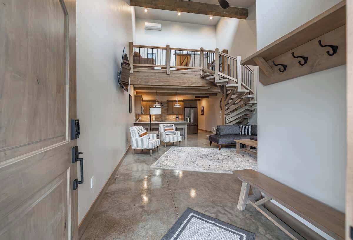 A spacious living area with a high ceiling, a wooden staircase, and a kitchen in the background.