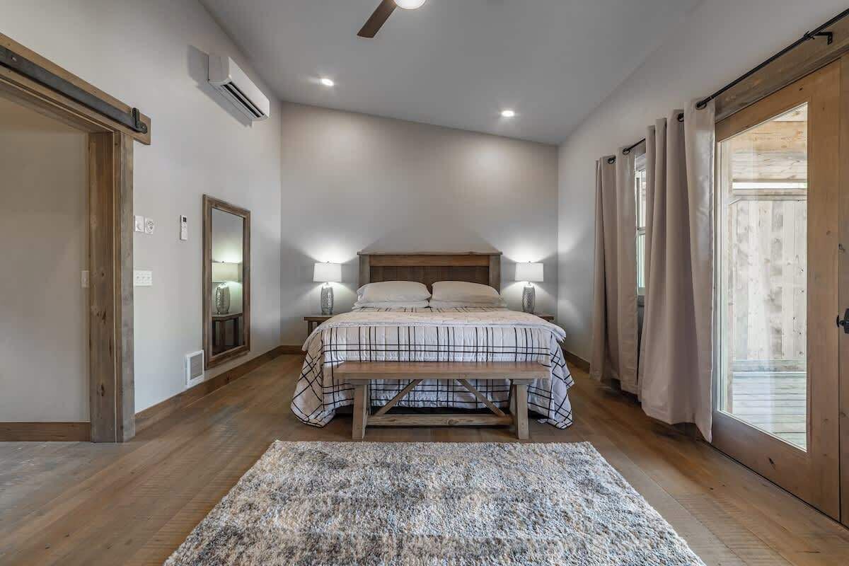 A bedroom with a bed, bedside lamps, a bench, a rug, and a large window with curtains.