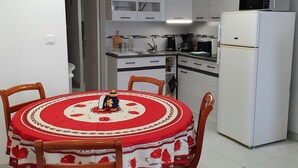 A compact kitchen with white cabinets, a microwave, and a refrigerator. A dining table with a red floral tablecloth and wooden chairs is in the foreground. - Whole house (Belvedere)