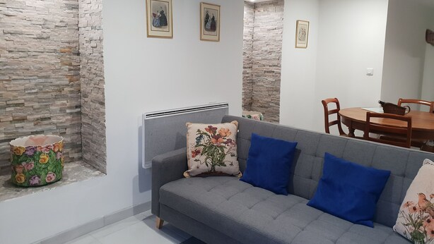 A living room with a grey sofa, blue cushions, and a floral-patterned side table. There are three framed pictures on the wall, a dining table with chairs, and a stone-textured wall. - Whole house (Belvedere)