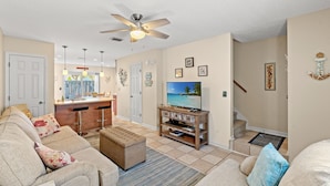 A living room with a sofa, ottoman, and a TV on a wooden stand. There is a ceiling fan, a kitchen area with a bar stool, and a staircase leading to another level. - 3 Min Walk to Beach-Perfect for Families-Pet Friendly (North Myrtle Beach)