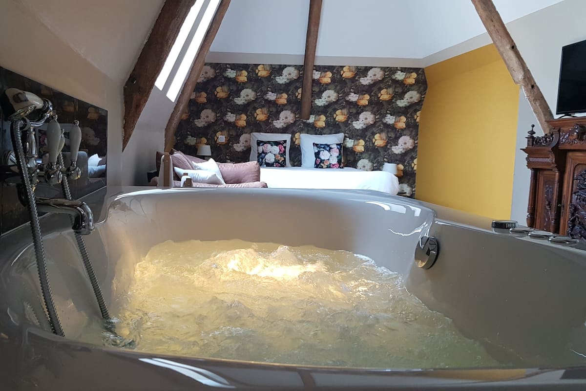 A room with a large bathtub, wooden beams, a floral wallpaper, and a bed with pillows.