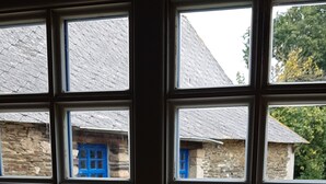 Vista a través de un ventanal que muestra un edificio de piedra con puertas y ventanas azules, y un jardín con plantas en macetas.