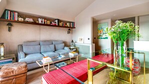 Un soggiorno moderno con un divano blu, un tavolino in vetro e un poggiapiedi rosso. Si nota una libreria piena di libri e oggetti decorativi, più un vaso con piante verdi su un tavolo in vetro.