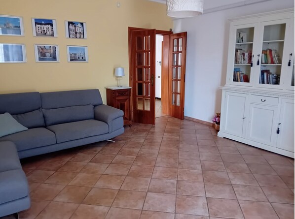 Een woonkamer met een bank, een kast vol boeken en een deur naar een andere kamer.