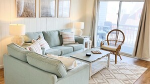 Ein Wohnzimmer mit einem hellgrünen Sofa, zwei Nachttischen mit Lampen, einem Couchtisch und einem Korbstuhl. An der Wand hängen drei gerahmte Bilder, ein großes Fenster mit Blick und ein gemustertes Teppich.
