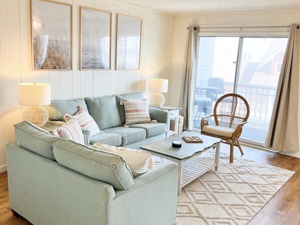 Un salon avec un canapé vert clair, deux tables d’appoint avec des lampes, une table basse et un fauteuil en osier. Trois tableaux encadrés ornent le mur, une grande fenêtre offre une vue dégagée et un tapis à motifs recouvre le sol.