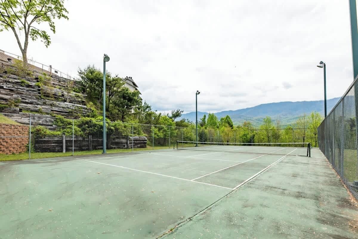 An outdoor tennis court with a net, surrounded by a fence, and a hillside with trees and a building in the background.