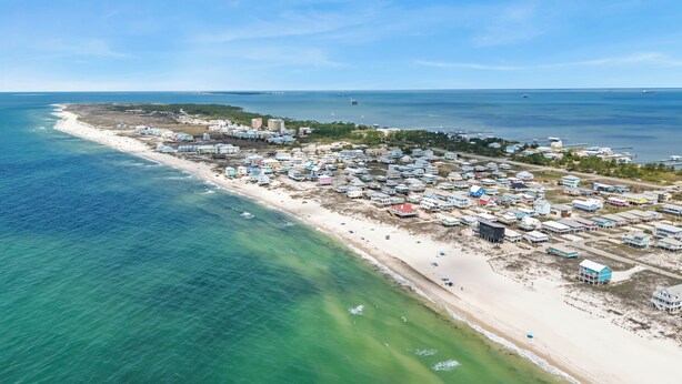 En vy över en kuststad från ovan, med bostäder som ligger direkt vid stranden, en sandstrand och en klarblå himmel.