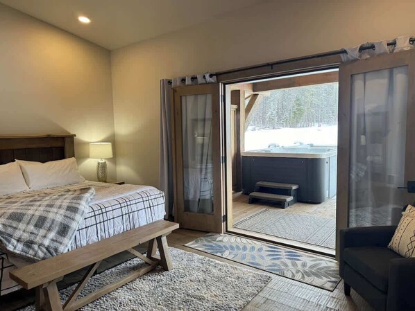 A bedroom with a bed, a bench, a nightstand, a lamp, a sofa, and a sliding glass door leading to a hot tub area. - Luxury Condo•Private Hot Tub•Mtn Views•7 mi to GNP (Coram)