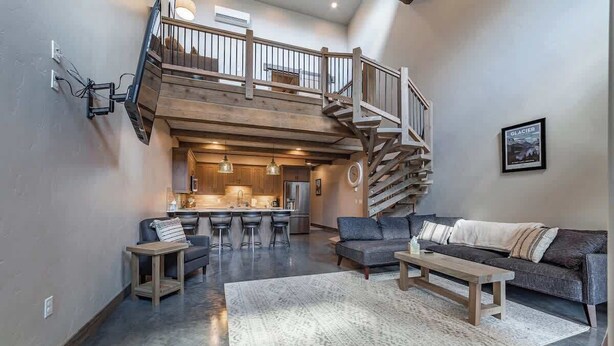 A spacious living area with a high ceiling, a wooden staircase, a kitchen in the background, and a large rug on the floor. - Luxury Condo•Private Hot Tub•Mtn Views•7 mi to GNP (Coram)