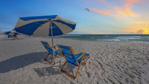A beach with a blue and white umbrella, two blue beach chairs, and a seagull flying in the sky.