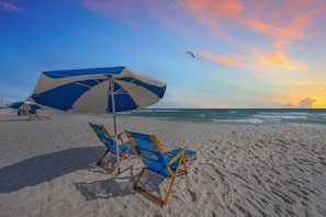 A beach with a blue and white umbrella, two blue beach chairs, and a seagull flying in the sky.