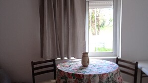 Een kamer met een ronde tafel, bedekt met een bloemrijk tafelkleed, twee stoelen, een vaas op de tafel, een raam met gordijnen en een bank tegen de muur.