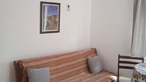 Een kamer met een gestreept bankje, een kleine tafel met een tafelkleed met bloemen, en een lijst met een afbeelding aan de muur.