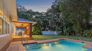A swimming pool with steps leading to a covered patio area, surrounded by lush greenery and trees. - Hyde Park Villa w/ Saltwater Pool + Fire Place! (Sarasota)