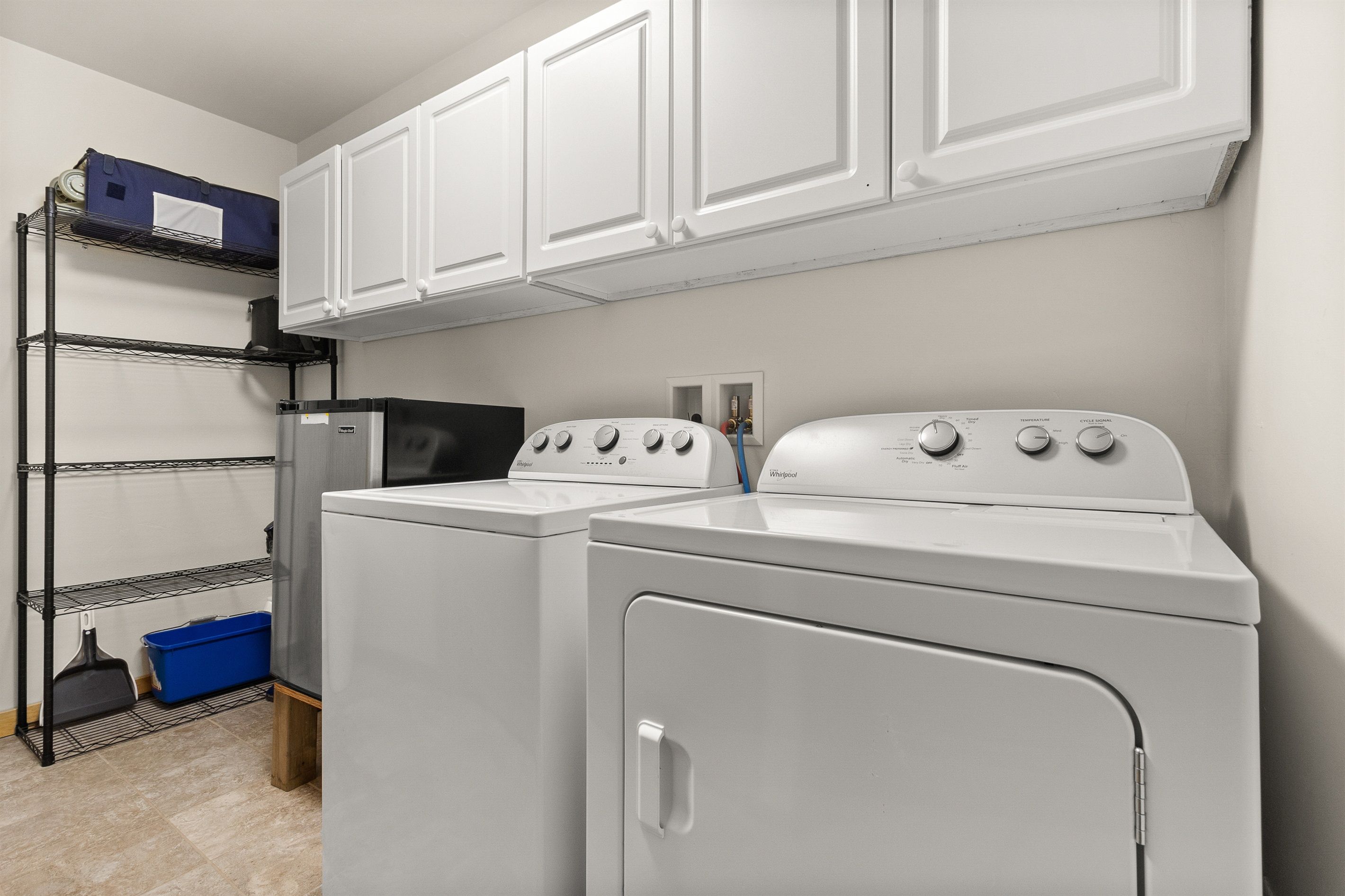 A laundry area with two white front-loading washers, a metal shelving unit with a blue bin, and white cabinets above.