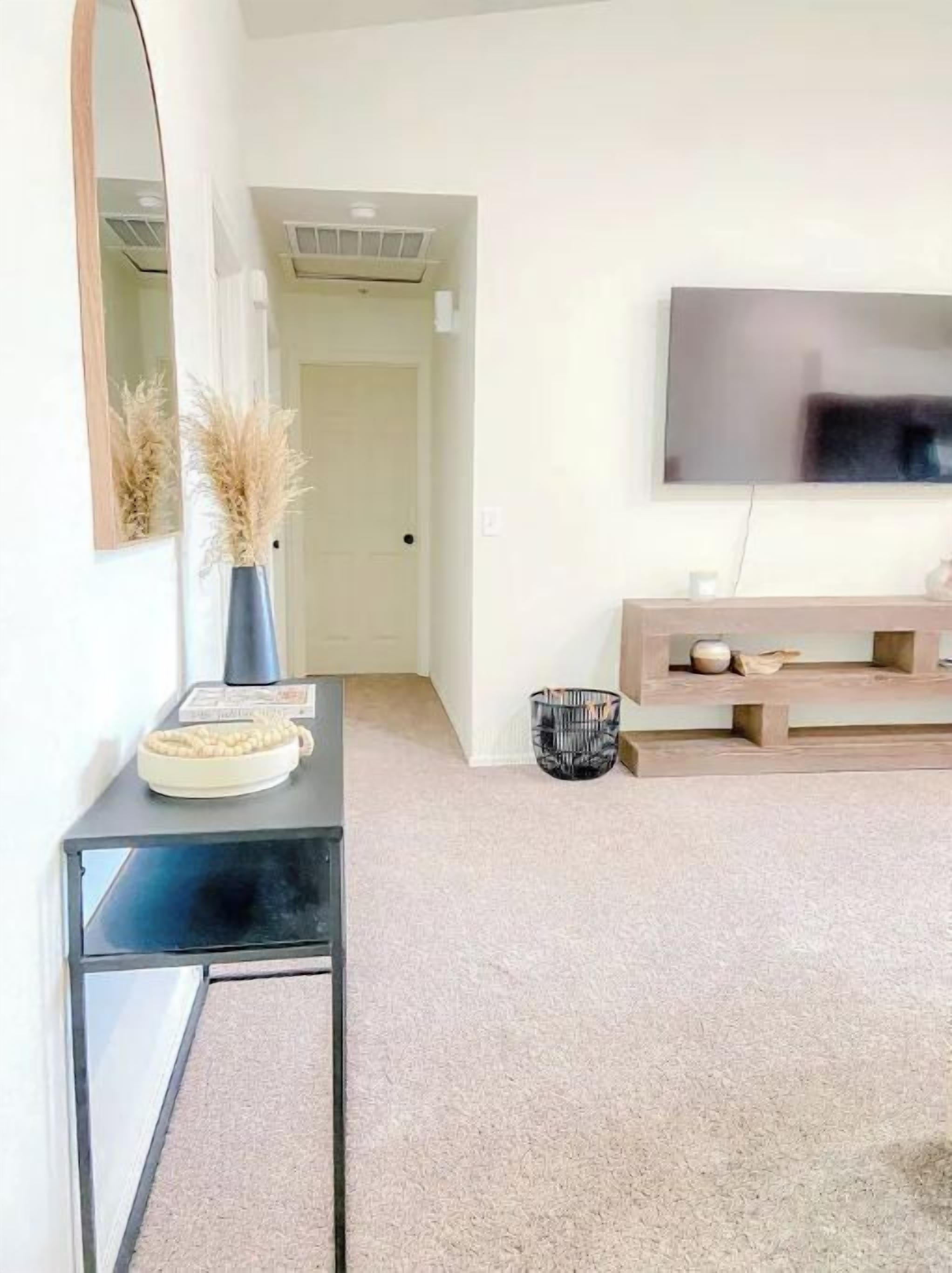 A modern living room with a black console table, a vase with dried plants, a flat-screen TV on a wooden stand, and a hallway leading to another room.