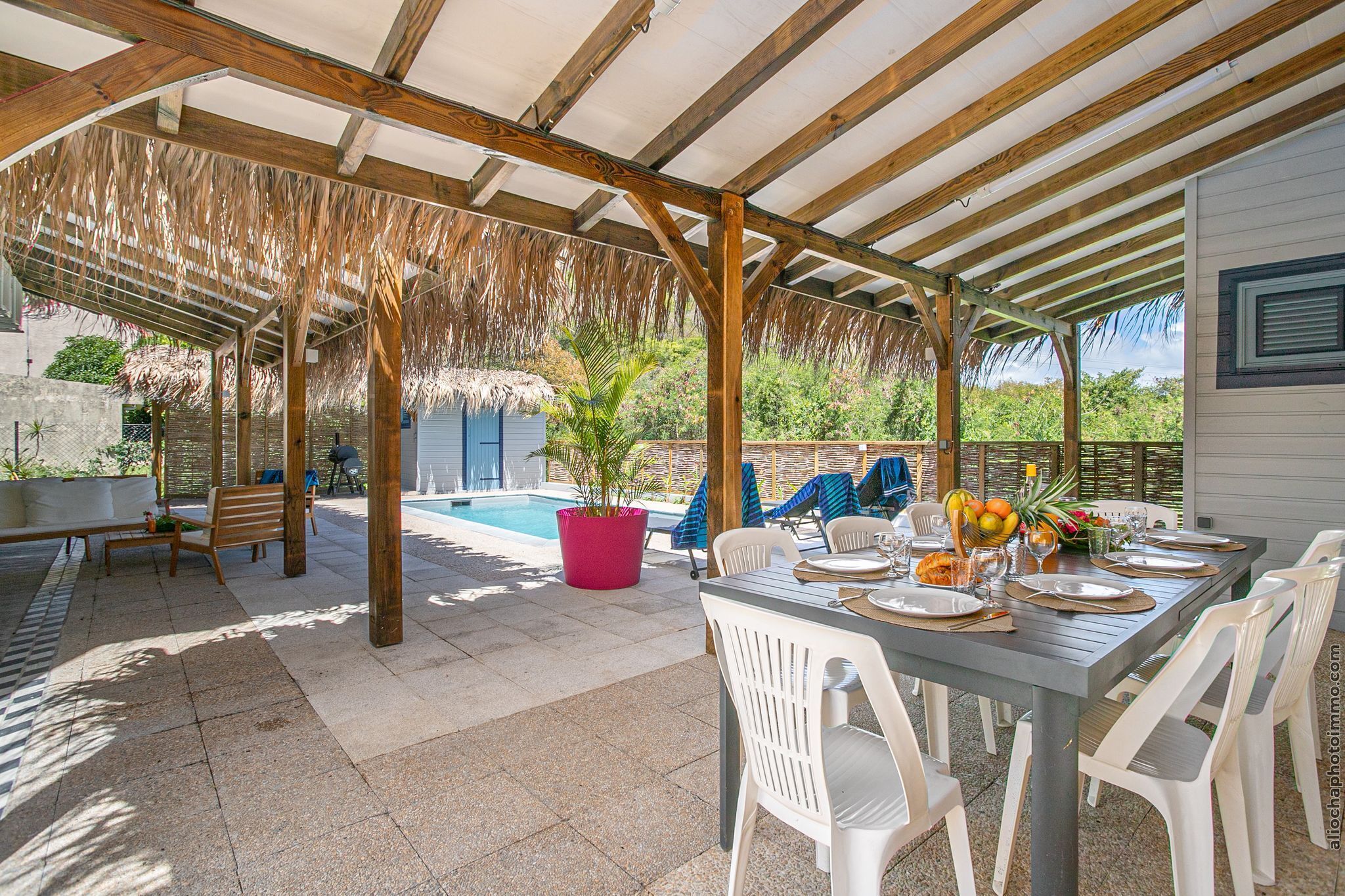 Een buitenterras met een zwembad, een houten pergola, zitplaatsen en een eettafel gedekt voor vier personen.