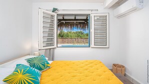 A bedroom with a bed, a window with open shutters, and a view of greenery outside. - Un séjour idéal à découvrir  (SAINTE ANNE)