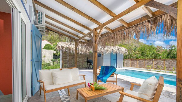 An outdoor patio with a wooden table, two white cushioned chairs, a pool, and a covered area with a thatched roof. - Un séjour idéal à découvrir  (SAINTE ANNE)