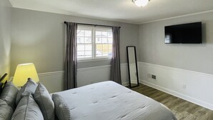 Un dormitorio con una cama, un televisor de pantalla plana montado en la pared, una ventana con cortinas y una lámpara de noche.