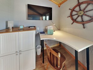 A compact room with a white desk, a mounted TV, a wooden chair, and a ship's wheel decoration on the wall.