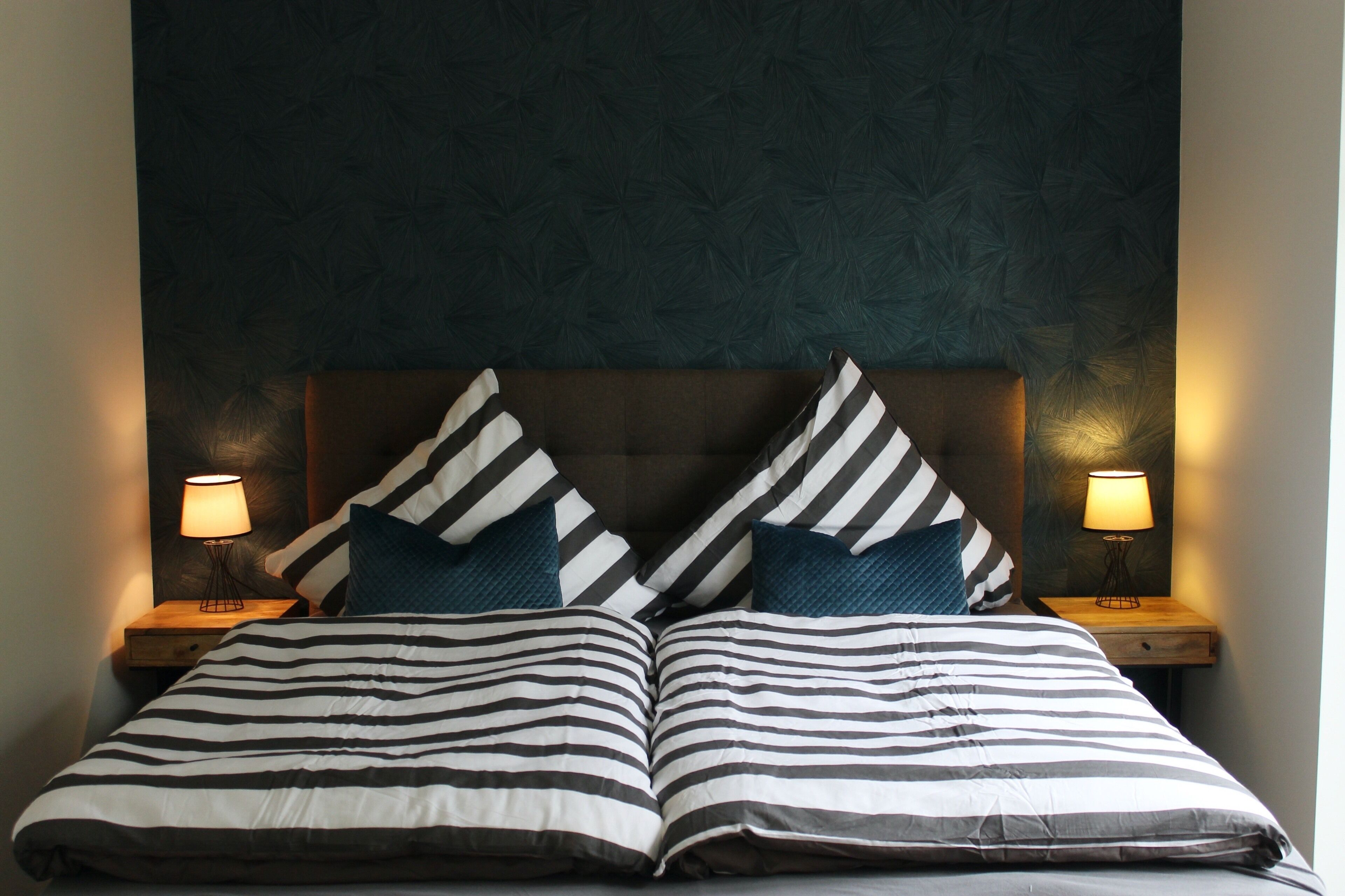 Ein Doppelbett mit gestreifter Bettwäsche und zwei Kissen, an dessen Seiten Nachttische mit Lampen stehen, in einem Zimmer mit dunkel gemustertem Wandpapier.