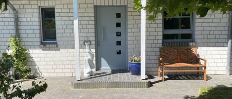 Un bâtiment à l’extérieur en briques blanches, avec une porte grise et un banc à l’extérieur.