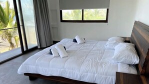 Una cama grande con ropa de cama blanca, cabecera de madera y vistas a palmeras a través de amplias ventanas.