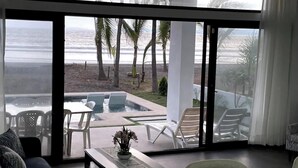 Una sala de estar moderna con un amplio ventanal corredera que da a una zona de piscina con sillones y vistas a la playa.