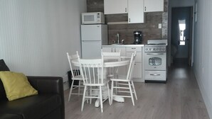 Een kleine keuken met witte kasten, een magnetron, koffiezetapparaat en een fornuis. Er is een eethoek met een witte tafel en stoelen. Een zwarte bank met een geel kussen staat op de voorgrond.