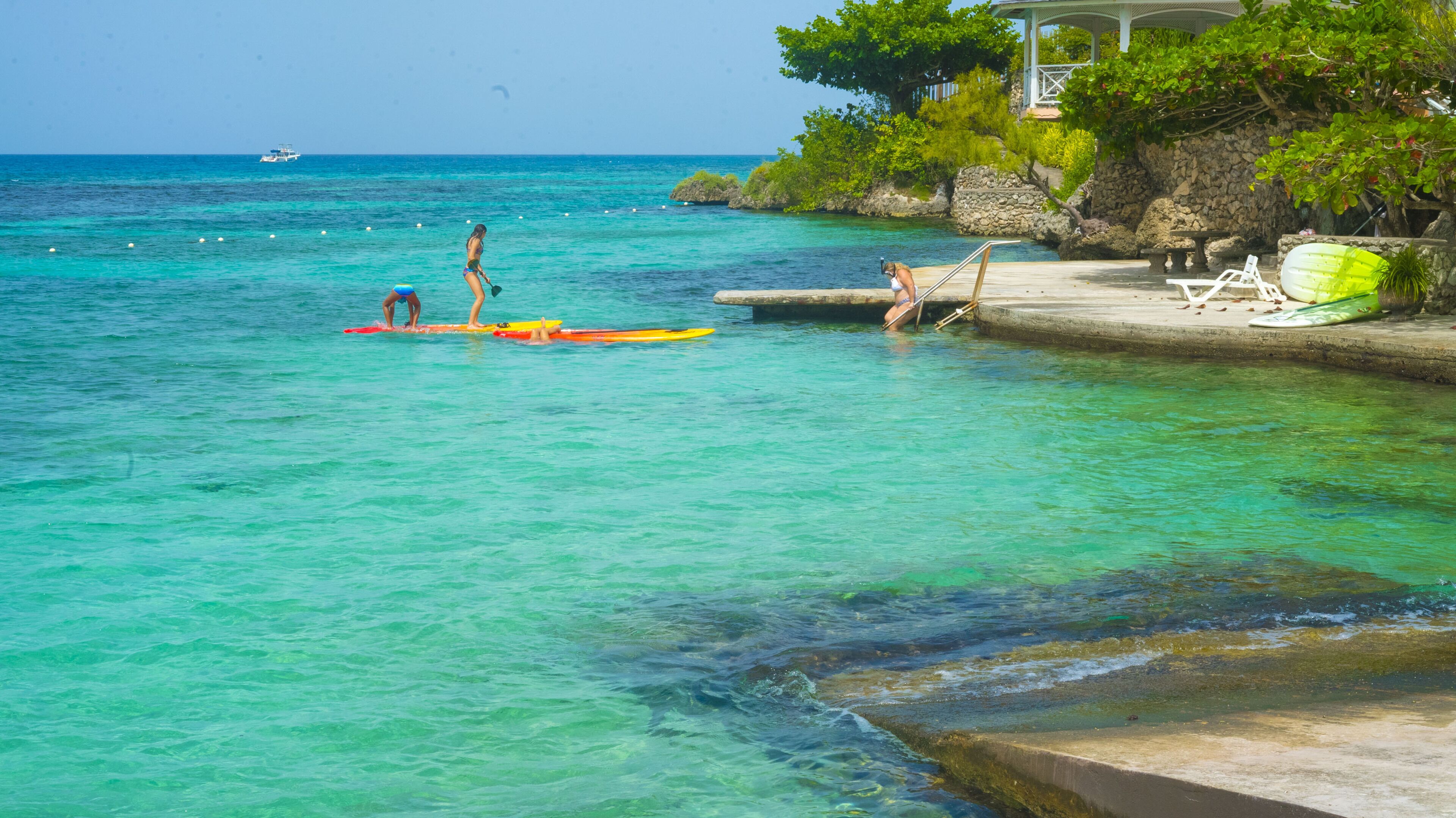 A clear turquoise sea with two people on surfboards, a gazebo, and a lounge chair by the water.