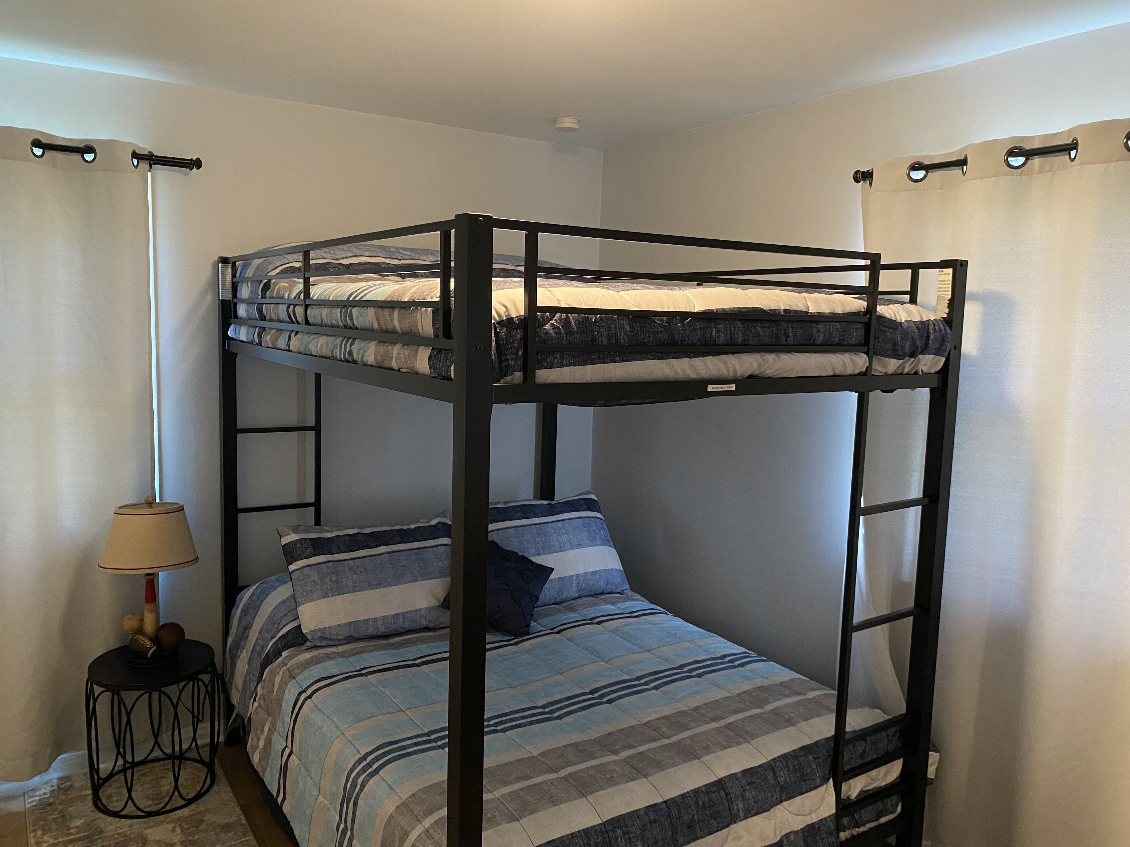 A bunk bed with a striped bedspread, a small round table with a lamp, and curtains on the windows.