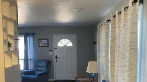 Una sala de estar con piso de madera, un sillón azul, una mesa de madera con una lámpara y una puerta con ventanal.