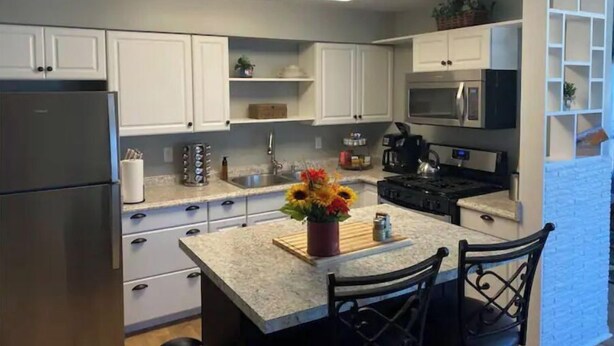 Una cocina moderna con armarios blancos, electrodomésticos de acero inoxidable, encimera de mármol y un pequeño comedor con dos sillas.