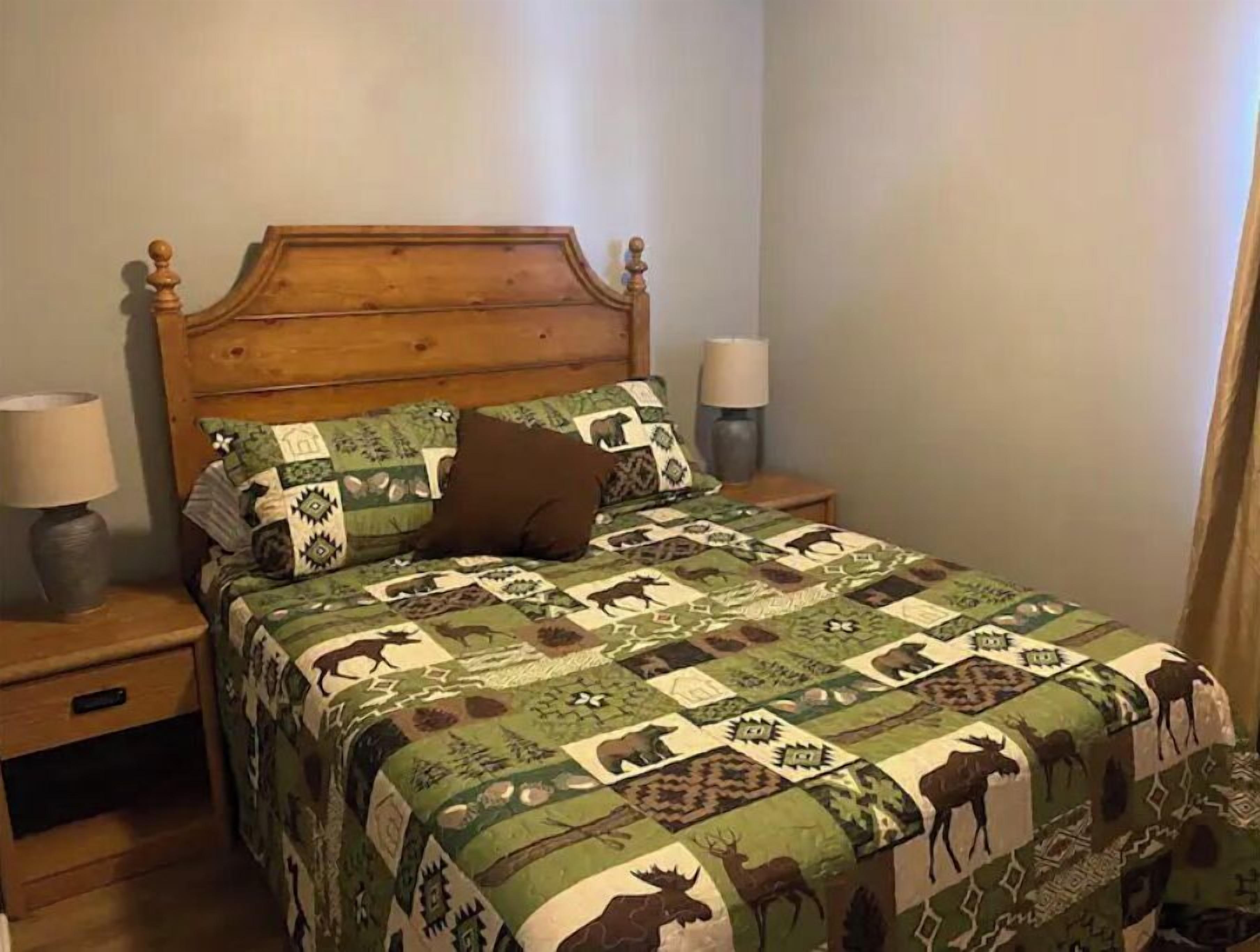 A bed with a wooden headboard, a quilt with animal patterns, two bedside lamps, and a nightstand with a lamp.