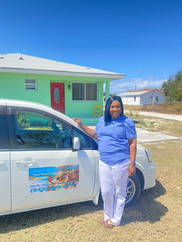 A person standing next to a white car parked in front of a green house with a red door.