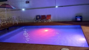 Uima-allas, jossa on sininen ja violetti valaistus, ja jonka ympärillä on valkoinen seinä sekä pieni patio, jossa on tuoleja ja aurinkovarjo.