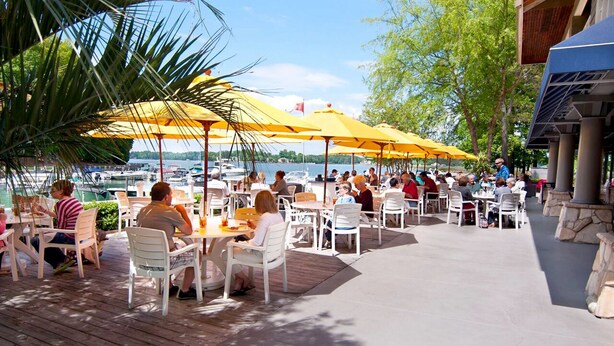 An outdoor dining area with tables and chairs under yellow umbrellas, overlooking a body of water with boats.