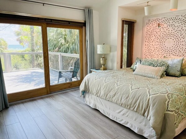 Un dormitorio con una cama grande, una mesita de noche con lámpara, un panel decorativo en la pared y un ventanal corredero que da a un espacio exterior con mesa y sillas.